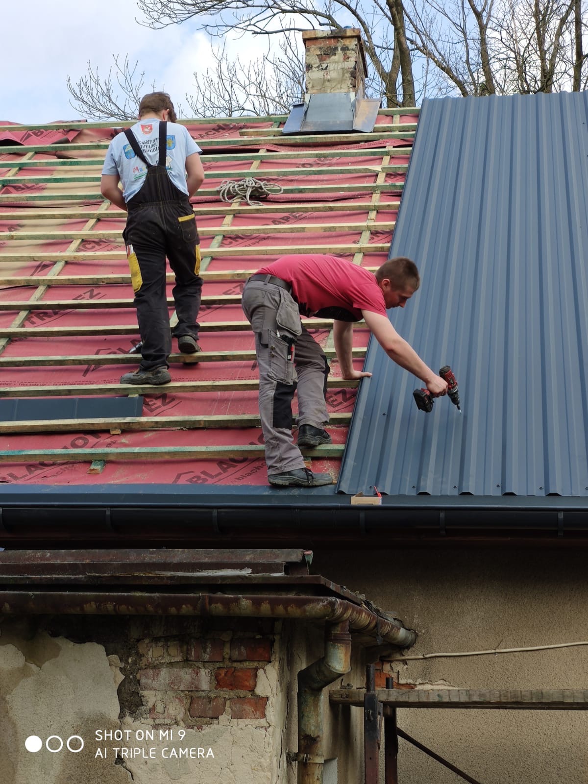 Dwóch dekarzy montuje blachodachówkę na drewnianej konstrukcji dachu; jeden przykręca wkrętarką arkusz blachy, drugi stoi w tle na łatach pokrytych czerwoną folią.