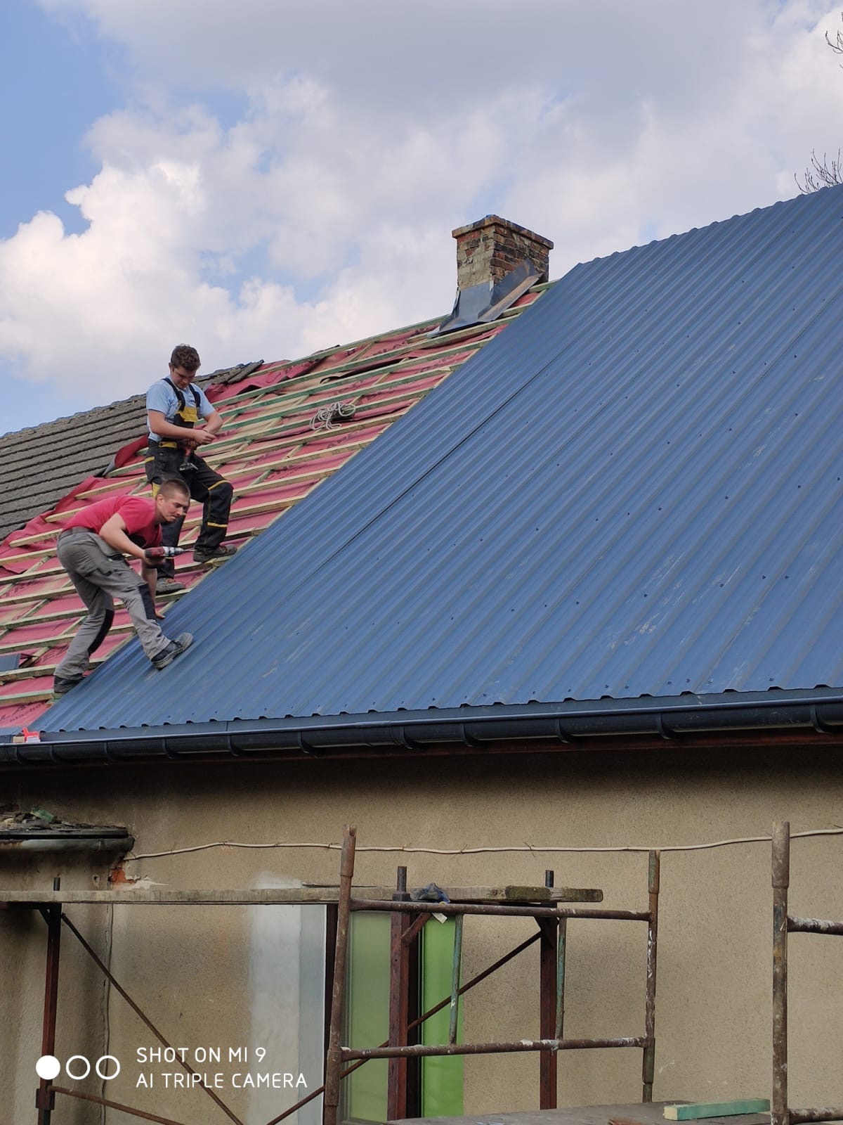 Dwóch dekarzy montuje blachodachówkę w kolorze grafitowym na połaci dachu, w tle widoczny komin z cegły i częściowo zdjęte stare pokrycie.