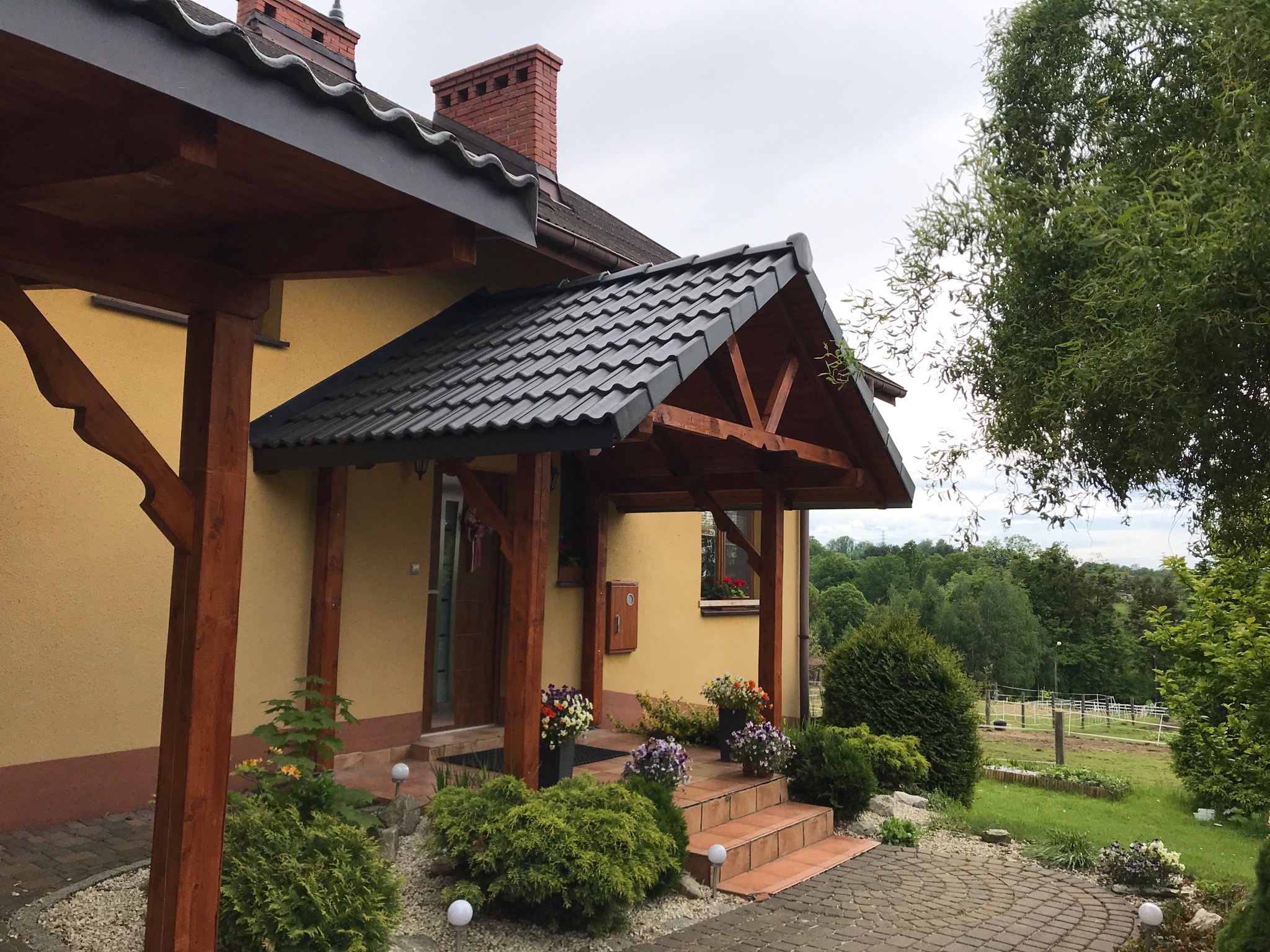 Żółty dom z ciemnym, spadzistym dachem i drewnianym zadaszeniem nad wejściem, ozdobiony kwiatami w donicach i zadbaną zielenią.