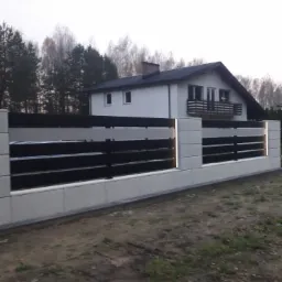 Nowoczesne ogrodzenie panelowe z betonowymi słupkami i poziomymi, ciemnymi przęsłami, oświetlone ledami, na tle domu jednorodzinnego i drzew.