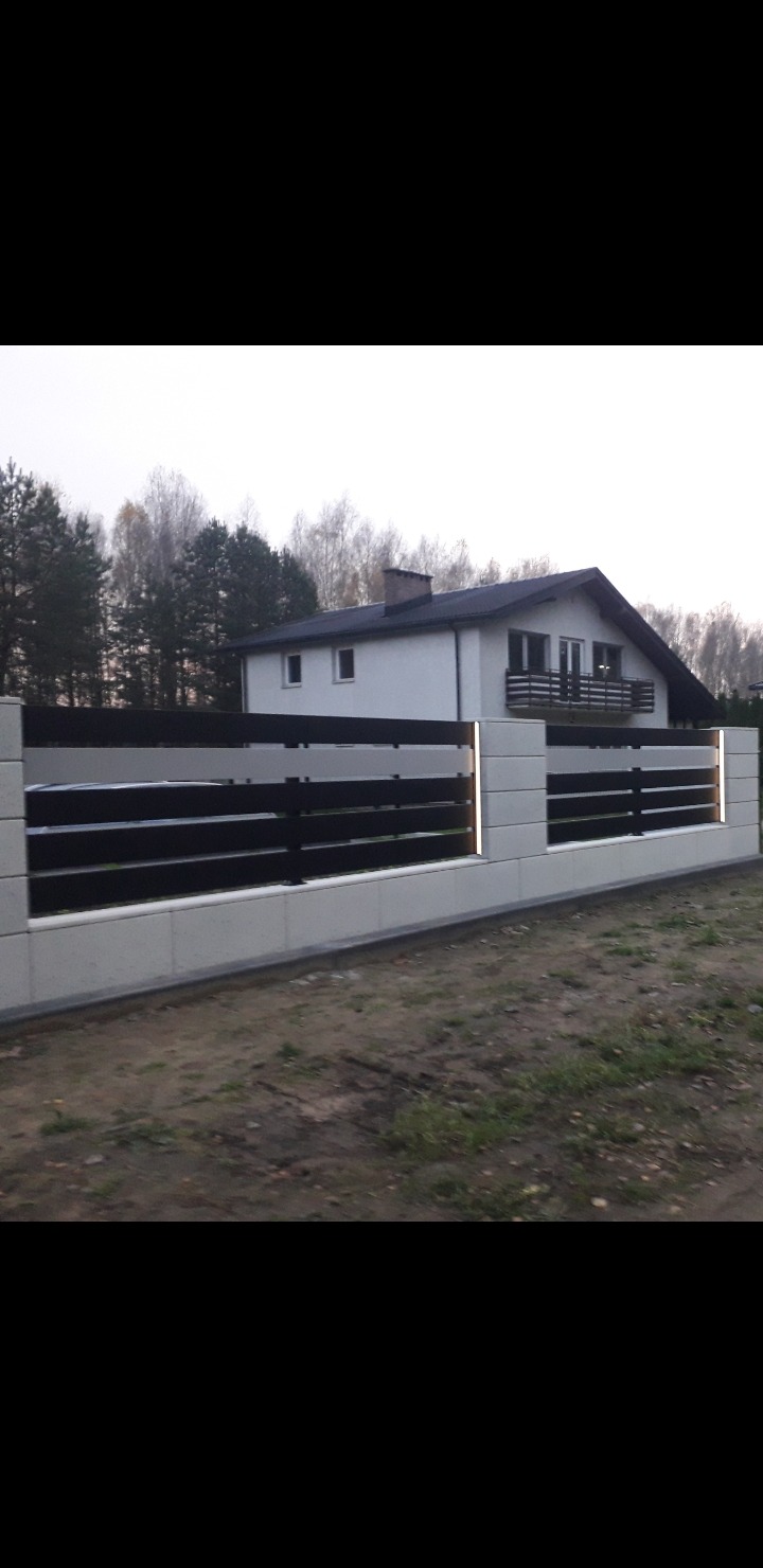 Nowoczesne ogrodzenie panelowe z betonowymi słupkami i poziomymi, ciemnymi przęsłami, oświetlone ledami, na tle domu jednorodzinnego i drzew.