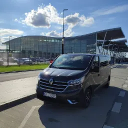 Ciemny van Renault z tablicą rejestracyjną OP 7705R zaparkowany przed nowoczesnym terminalem lotniska w słoneczny dzień.