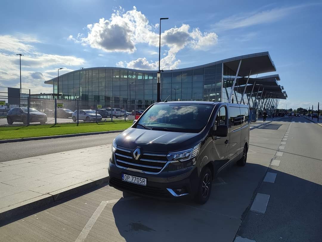 Ciemny van Renault z tablicą rejestracyjną OP 7705R zaparkowany przed nowoczesnym terminalem lotniska w słoneczny dzień.