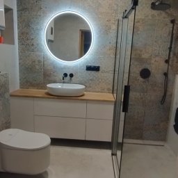 Paweł Lewoń - Nowoczesna łazienka z drewnianym sufitem, okrągłym lustrem z podświetleniem LED, białą umywalką nablatową, wiszącą toaletą i kabiną prysznicową z czarnymi detalami.