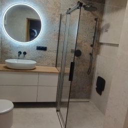 Paweł Lewoń - Nowoczesna łazienka z drewnianym stropem, okrągłym lustrem z podświetleniem LED, kabiną prysznicową typu walk-in z czarnymi detalami i umywalką nablatową.