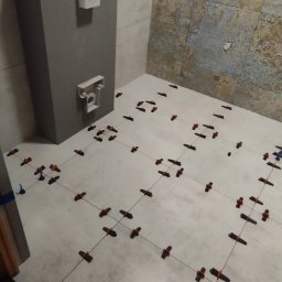 Paweł Lewoń - Białe płytki podłogowe w trakcie układania w łazience, widoczne krzyżyki dystansowe i sznurki poziomujące, z elementami instalacji sanitarnych na ścianie.