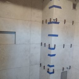 Paweł Lewoń - Ściana łazienki w trakcie wykańczania, widoczne płytki mocowane za pomocą klipsów i taśmy malarskiej, surowy beton na podłodze, drewniana belka pod sufitem.