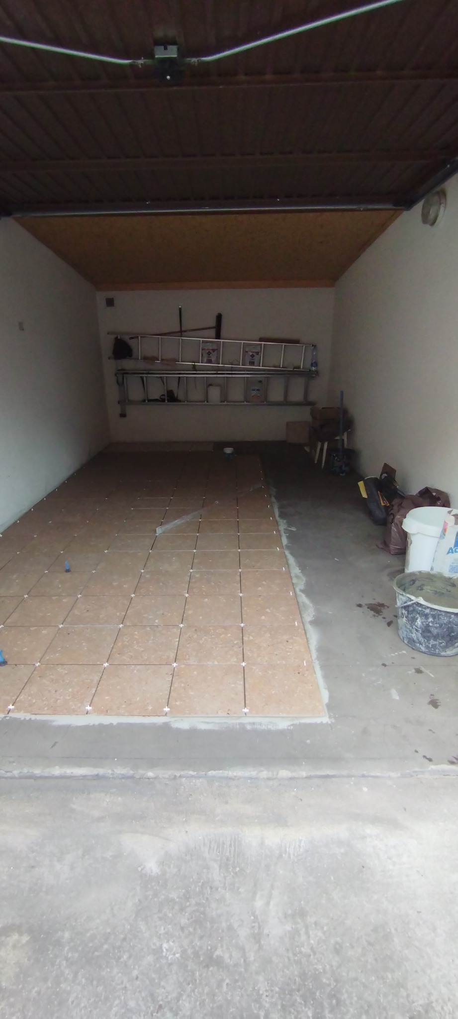 Proces układania brązowych płytek podłogowych w garażu, widoczne krzyżyki dystansowe, częściowo wykończona powierzchnia, drabiny oparta o ścianę w tle.