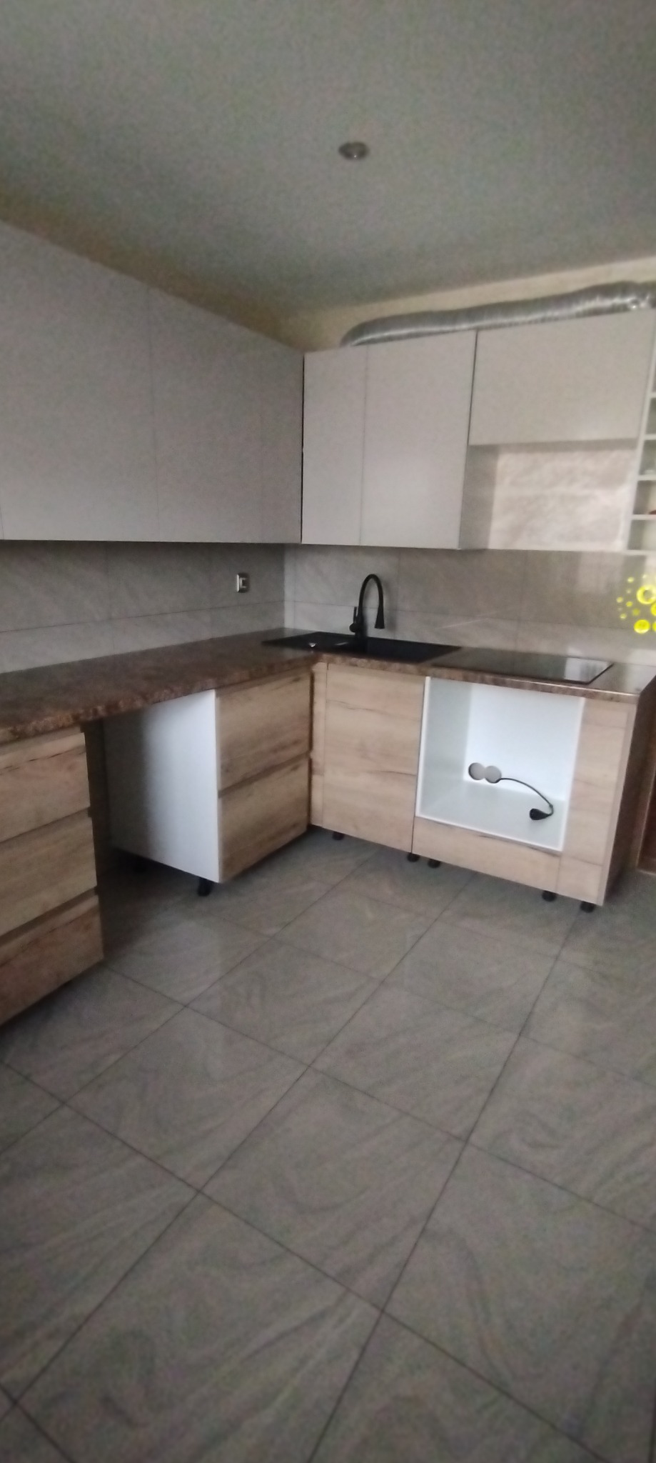 Częściowo zamontowana, narożna zabudowa kuchenna z jasnymi, drewnopodobnymi frontami, brązowym blatem i czarnym zlewem; widoczne niedokończone podłączenia elektryczne.