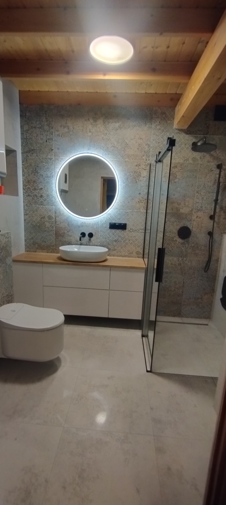 Nowoczesna łazienka z drewnianym sufitem, okrągłym lustrem z podświetleniem LED, białą umywalką nablatową, wiszącą toaletą i kabiną prysznicową z czarnymi detalami.