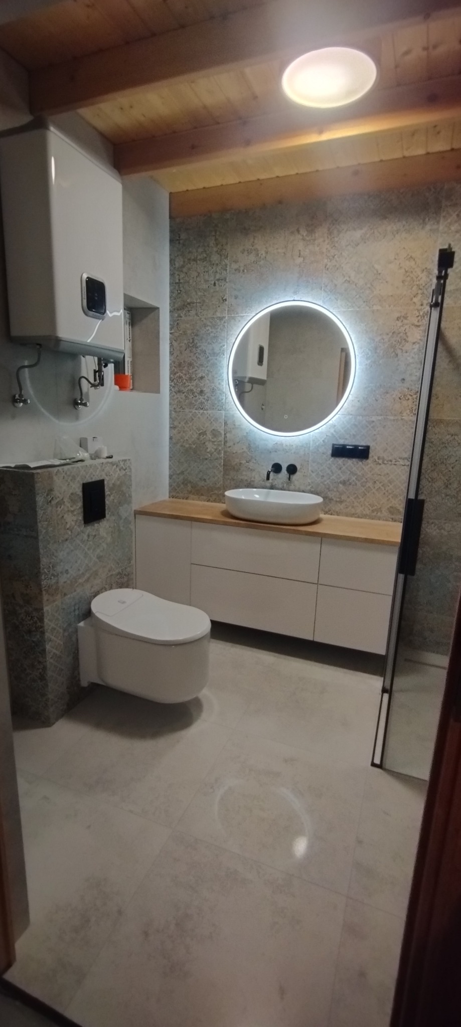 Nowoczesna łazienka z podwieszaną toaletą, umywalką nablatową na drewnianym blacie i okrągłym lustrem z podświetleniem LED, wykończona płytkami w odcieniach szarości i beżu, z widocznym bojlerem...