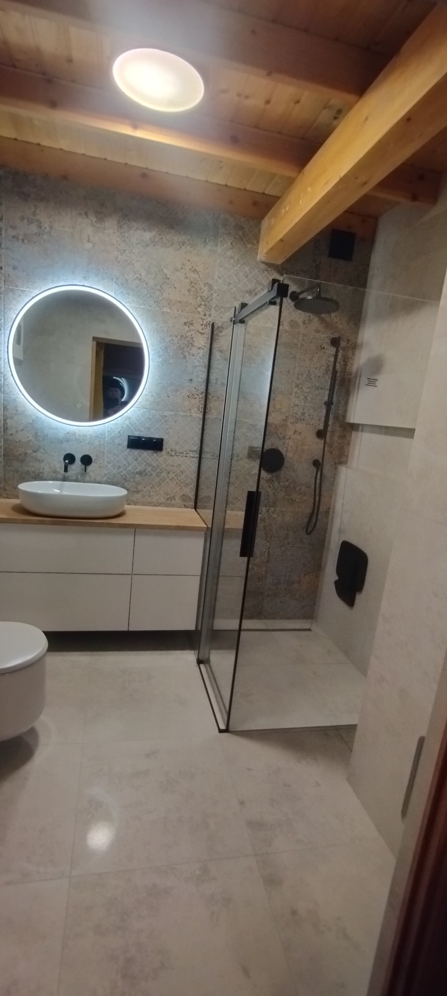 Nowoczesna łazienka z drewnianym stropem, okrągłym lustrem z podświetleniem LED, kabiną prysznicową typu walk-in z czarnymi detalami i umywalką nablatową.