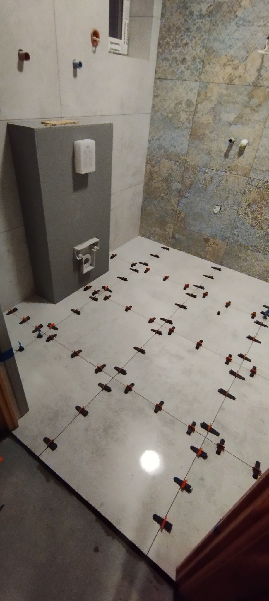 Białe płytki podłogowe w trakcie układania w łazience, widoczne krzyżyki dystansowe i sznurki poziomujące, z elementami instalacji sanitarnych na ścianie.