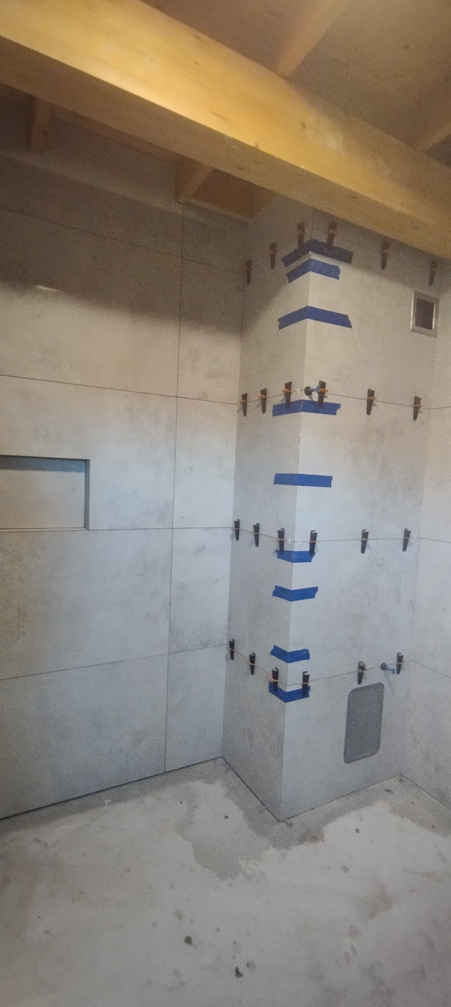 Ściana łazienki w trakcie wykańczania, widoczne płytki mocowane za pomocą klipsów i taśmy malarskiej, surowy beton na podłodze, drewniana belka pod sufitem.