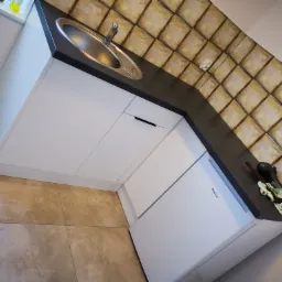 Kątowe ujęcie białych szafek kuchennych z czarnym blatem i okrągłym zlewem, na blacie leżą ściereczki obok wbudowanej lodówki Amica, w tle kwadratowe płytki na ścianie.