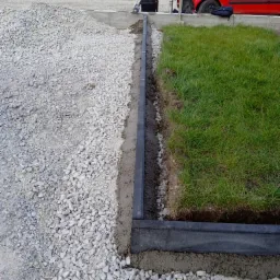 Przygotowanie podłoża pod trawnik: krawężnik z tworzywa sztucznego oddziela warstwę żwiru od trawy, widoczny świeży beton i sterta drewna w tle.