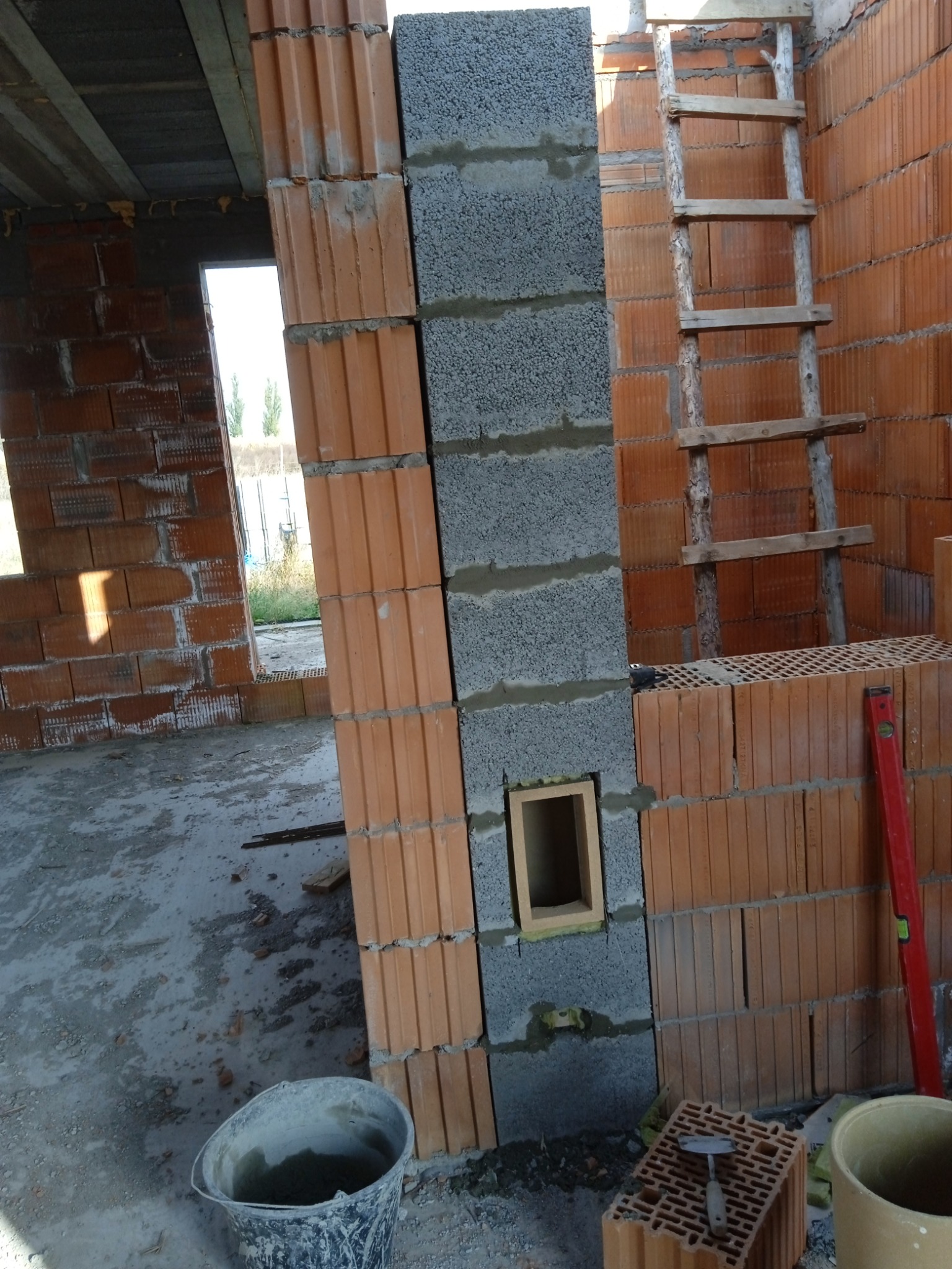 Wznoszenie ściany z cegieł ceramicznych i bloczków betonowych, widoczna drabina drewniana, poziomica i wiadro z zaprawą.