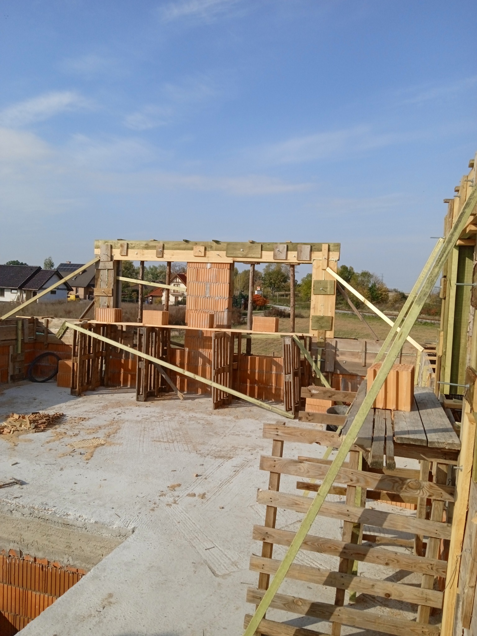 Budowa domu jednorodzinnego: wznoszenie ścian z pustaków ceramicznych Porotherm, widoczne drewniane szalunki i podpory, etap murowania pierwszej kondygnacji, błękitne niebo w tle.