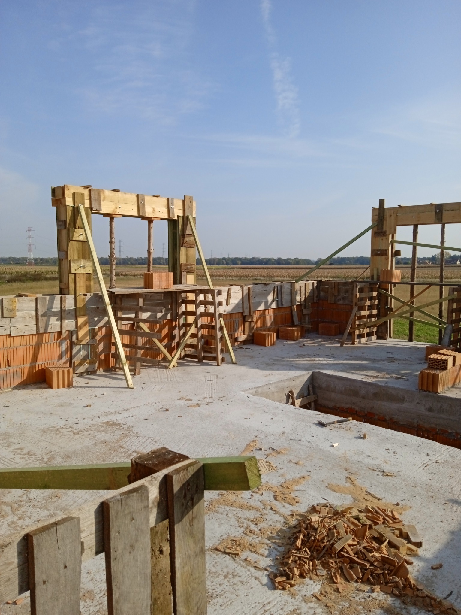Budowa domu w trakcie wznoszenia ścian z cegły i drewnianych szalunków, widok na plac budowy z rozrzuconymi fragmentami drewna.