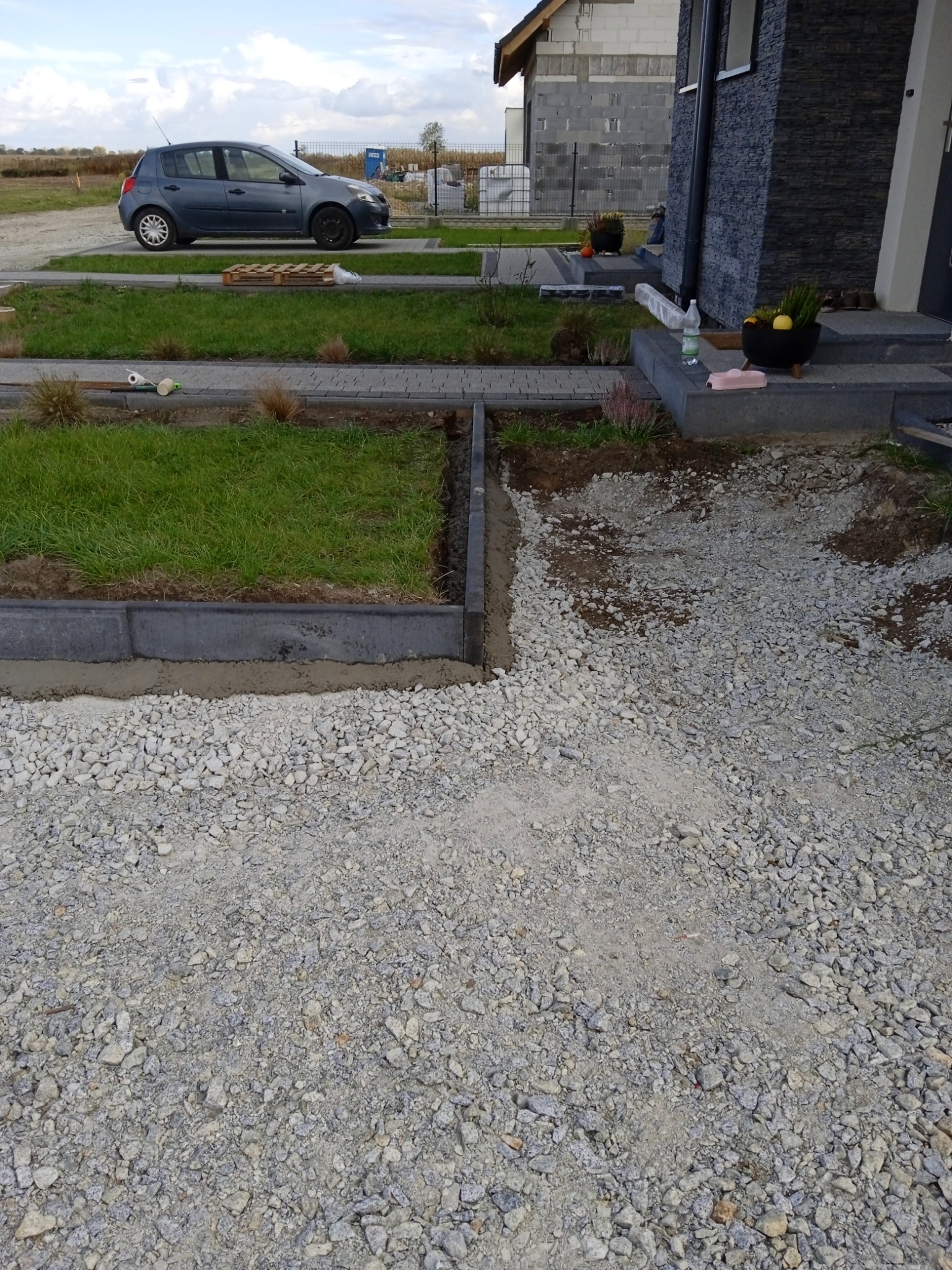 Przygotowanie terenu pod trawnik: ułożone obrzeża z kostki betonowej, podsypka z drobnego kruszywa, widoczne ślady cementu, w tle dom w trakcie budowy i zaparkowany samochód.