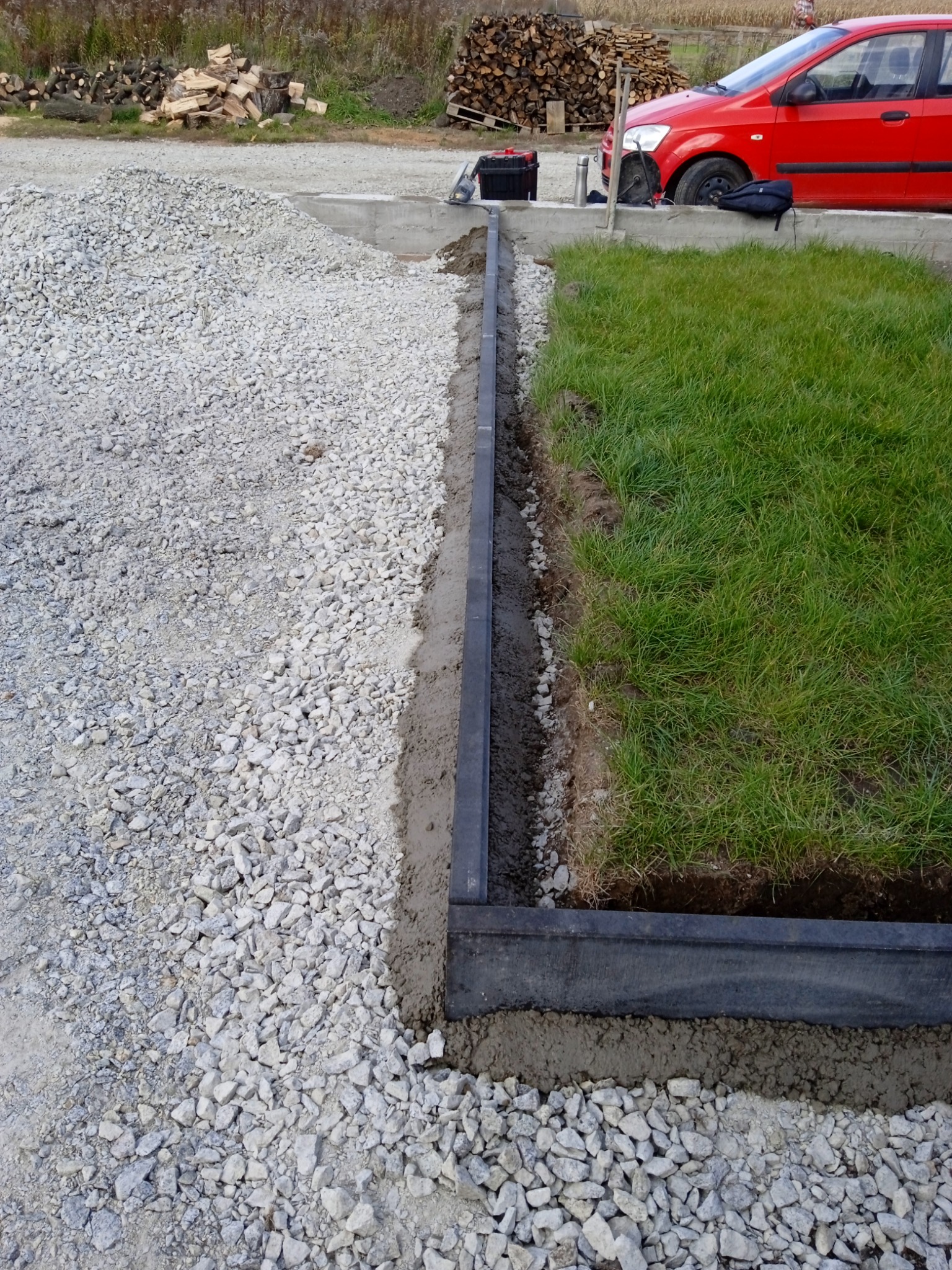 Przygotowanie podłoża pod trawnik: krawężnik z tworzywa sztucznego oddziela warstwę żwiru od trawy, widoczny świeży beton i sterta drewna w tle.