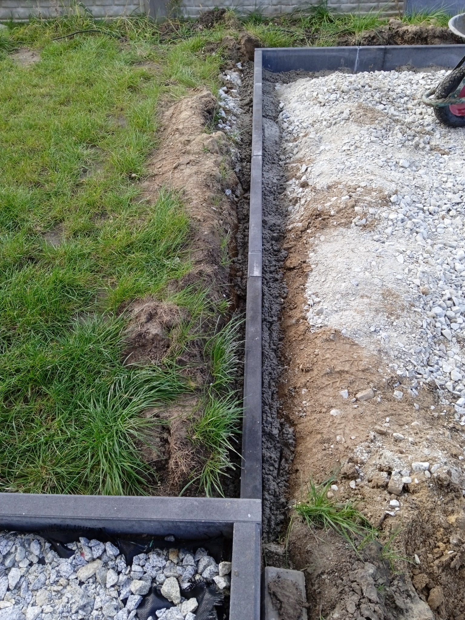 Przygotowanie terenu pod ścieżkę: ułożone obrzeża z kostki betonowej, wykopana ziemia, wysypany żwir, widoczny trawnik i fragment taczki.