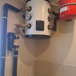 Biały zbiornik akumulacyjny Auraton z czarnymi rurami, czerwonym zbiornikiem wyrównawczym i filtrem magnetycznym Adey, zamontowany w rogu pomieszczenia wyłożonego płytkami.