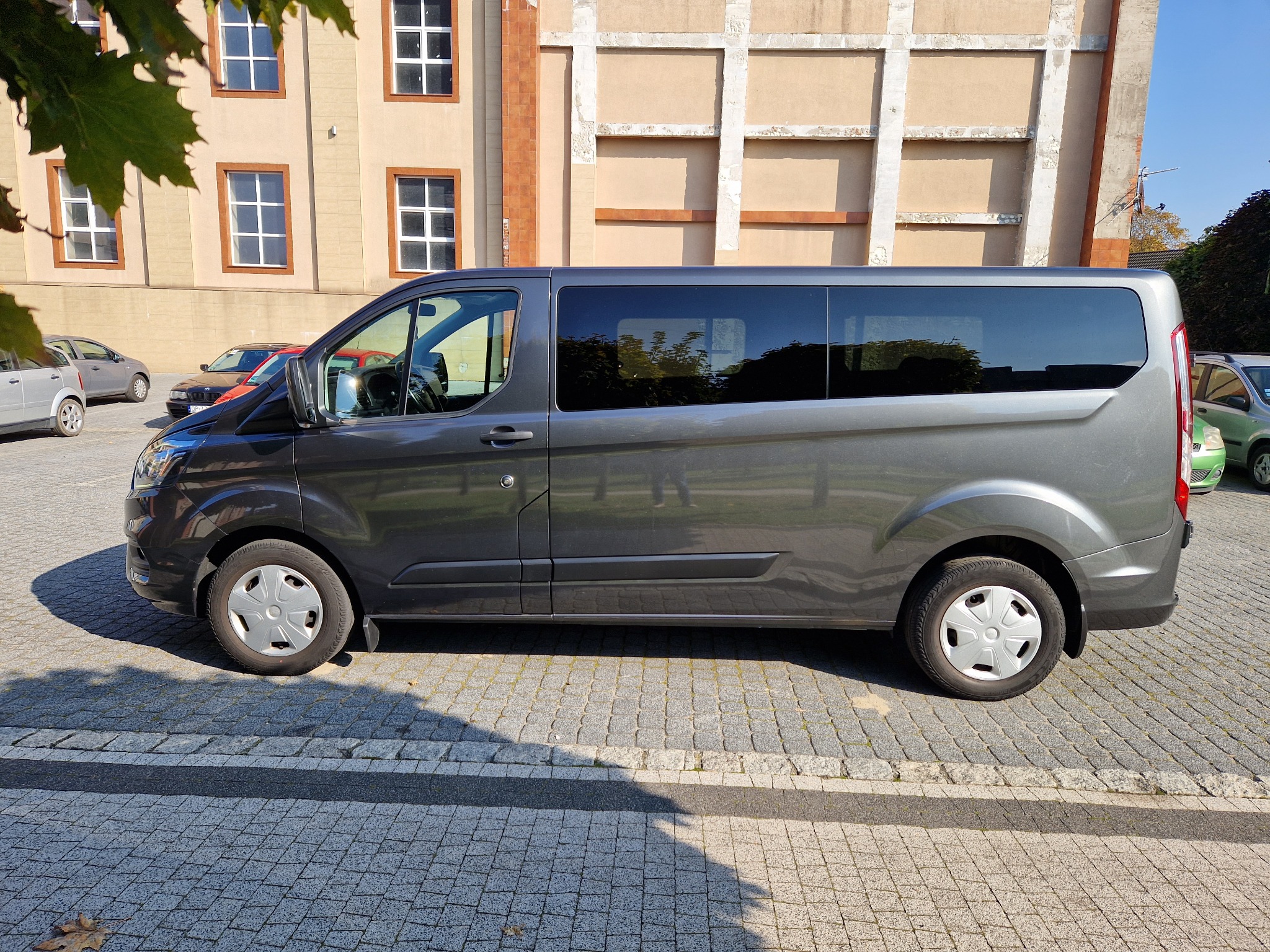 Szary van zaparkowany na brukowanym parkingu przed budynkiem z beżową fasadą i brązowymi akcentami.