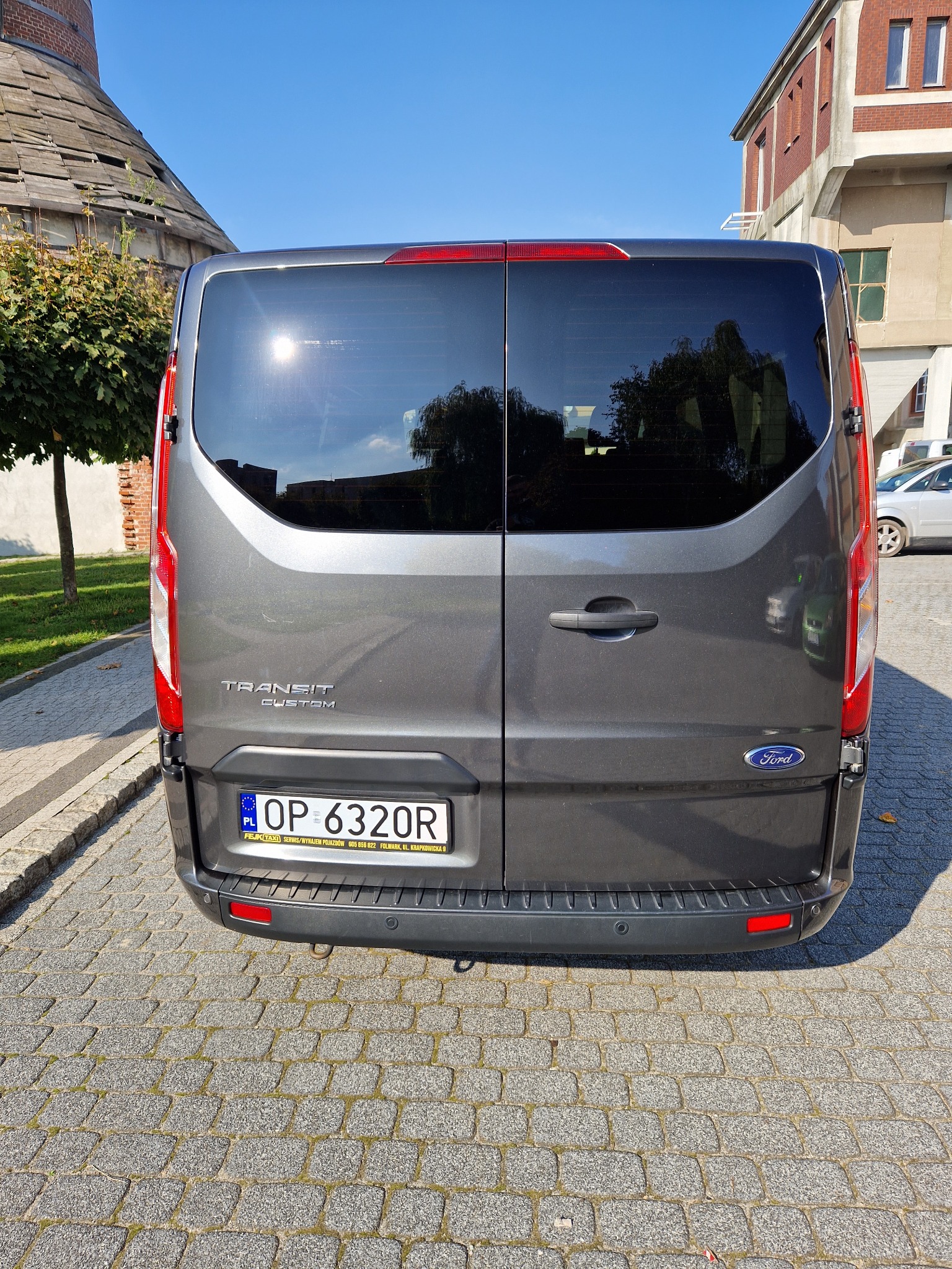 Tył szarego Forda Transit Custom z polską tablicą rejestracyjną OP 6320R, na tle ceglanego budynku i brukowanej drogi. Na tablicy widoczna nazwa firmy 'FEJK TAXI SERWIS/WYNAJEM POJAZDÓW'.