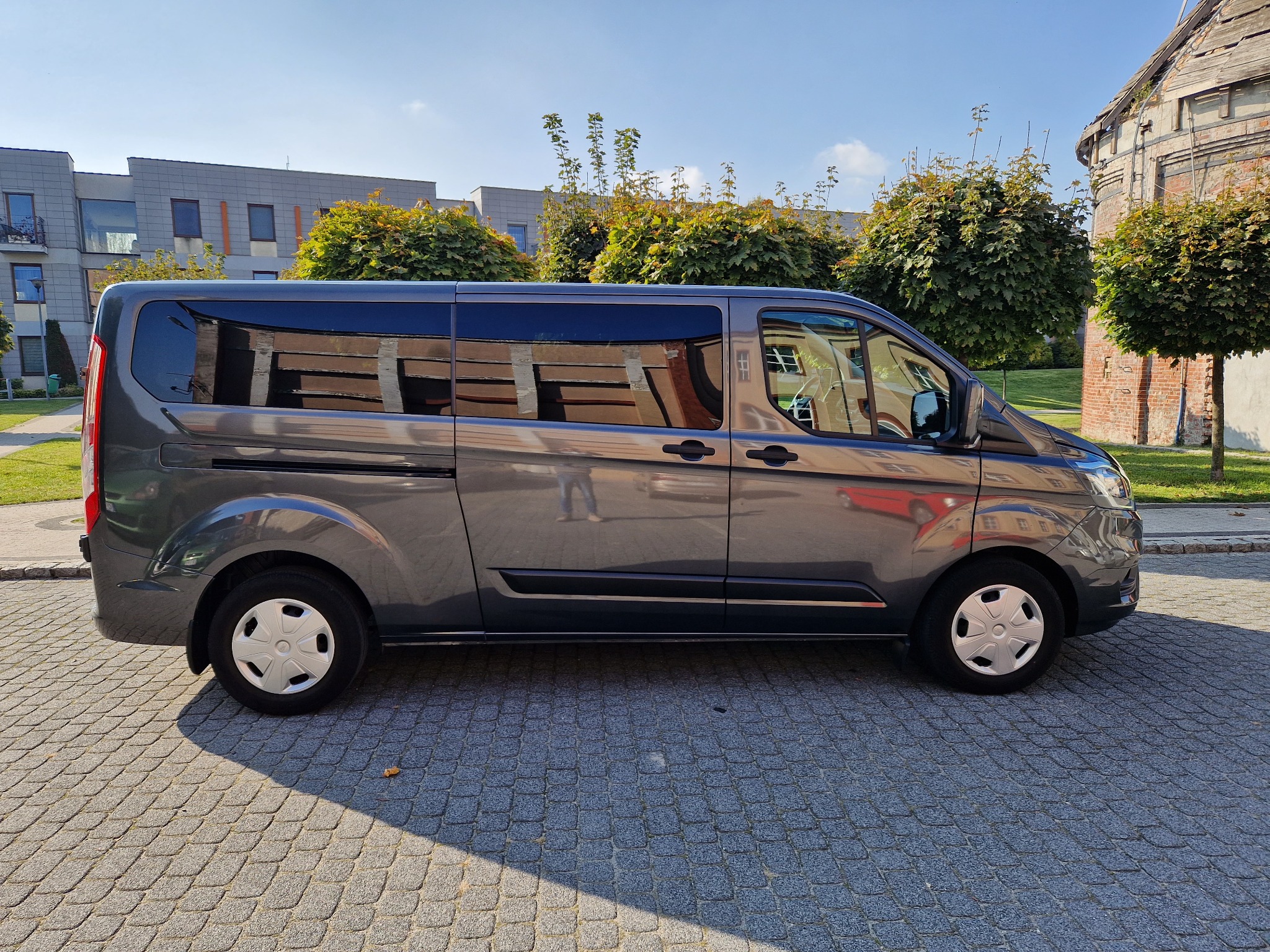 Szary van pasażerski Ford Transit Custom w ujęciu z boku, zaparkowany na brukowanej ulicy w słoneczny dzień, z budynkami i zielenią w tle.