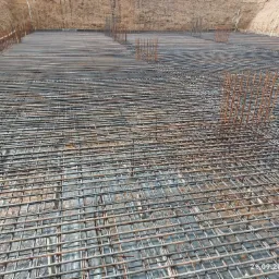 Zbrojenie fundamentu płyty, widoczne siatki z prętów stalowych ułożone na folii izolacyjnej, pionowe pręty wystające w miejscach przyszłych słupów, wykop ziemny w tle.