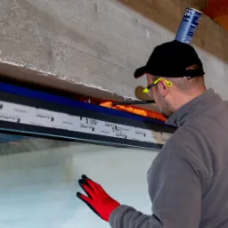 Pracownik w okularach ochronnych uszczelnia pianką montażową ramę okienną z profili aluminiowych marki Aluprof; puszka z pianką umieszczona na czapce dla wygody.