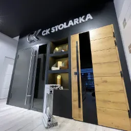 Ekspozycja w salonie stolarki: przekrój ramy okiennej, drzwi wejściowe z drewnopodobnym dekorem, szare drzwi z długim uchwytem i białe drzwi z poziomymi wgłębieniami, podświetlone logo 'CK Stolarka'.