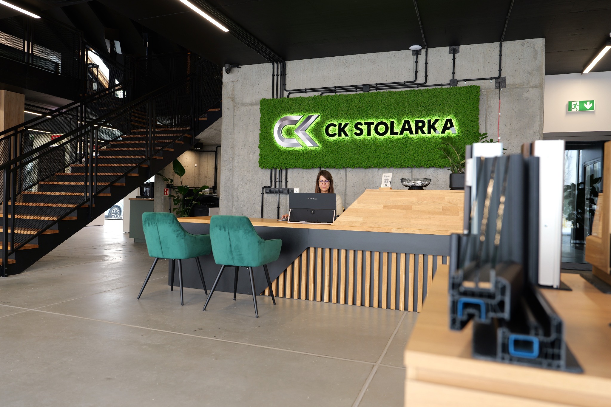 Nowoczesne biuro firmy CK Stolarka z recepcją, zielonym logo na ścianie i próbkami profili okiennych na pierwszym planie. Widoczne schody i pracownik przy laptopie.