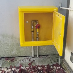 GTA Gaztechnika - Żółta skrzynka z otwartymi drzwiczkami, zawierająca instalację gazową z reduktorem i zaworami, umieszczona na zewnątrz budynku, otoczona kamieniami i liśćmi.