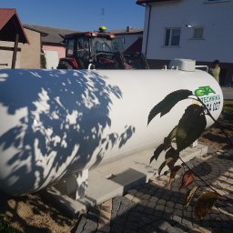 GTA Gaztechnika - Biały zbiornik na gaz LPG z logo firmy, zamontowany na betonowej podstawie na posesji, w tle traktor i budynek mieszkalny.