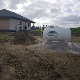GTA Gaztechnika - Montaż naziemnego zbiornika na gaz płynny przy nowo budowanym domu jednorodzinnym, widoczny wykop pod instalację, logo firmy Gaztechnika i numer telefonu na zbiorniku.