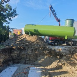 GTA Gaztechnika - Montaż podziemnego zbiornika na gaz płynny za pomocą dźwigu, widoczny wykop z betonowymi płytami i pomarańczowymi rurami, ciężarówka z drugim zbiornikiem w tle.