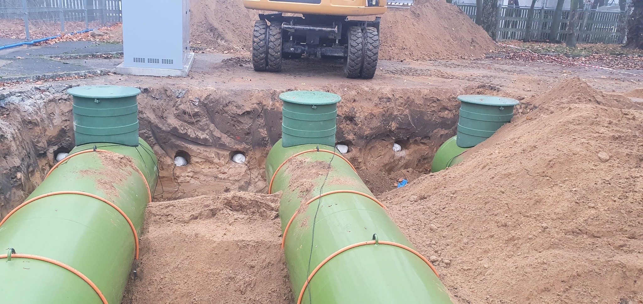 Instalacja zbiorników gazowych w wykopie, widoczne zielone zbiorniki z pomarańczowymi pierścieniami, ziemia i koparka w tle.