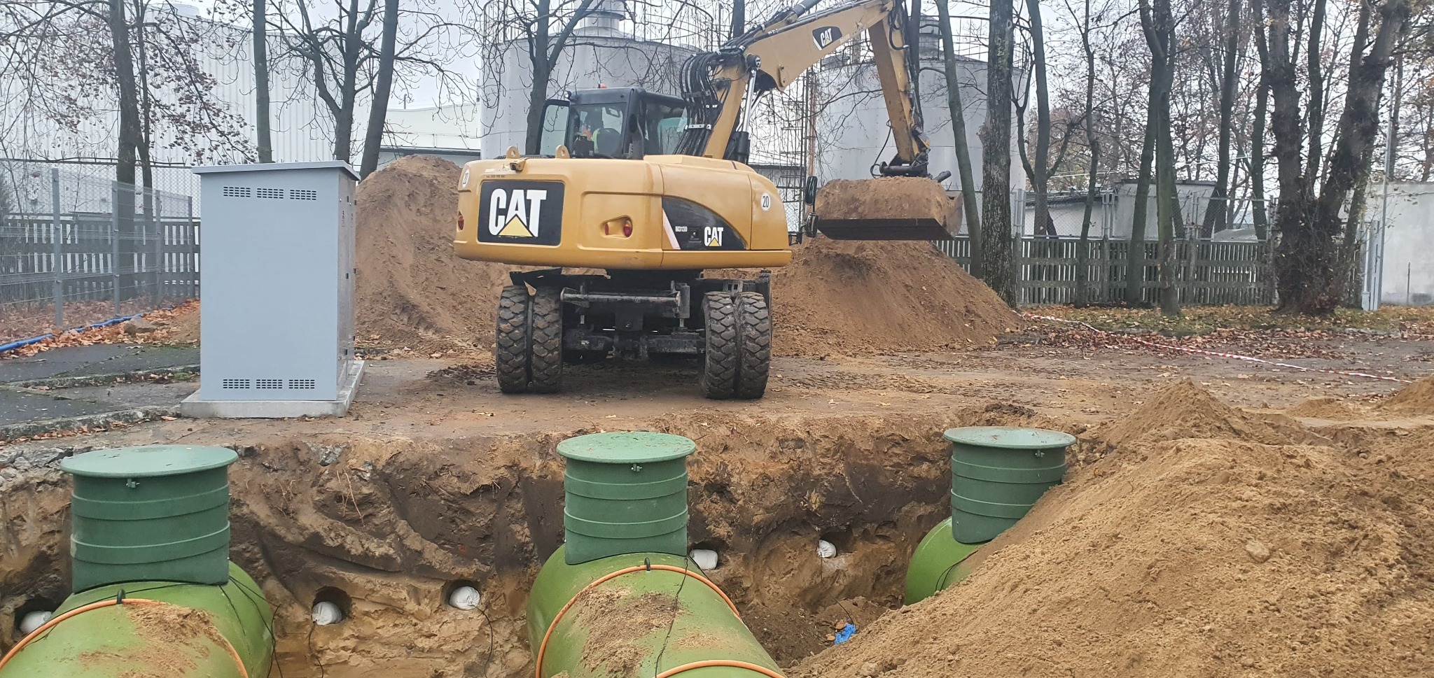 Żółta koparka CAT zasypuje wykop z zielonymi zbiornikami z białymi rurami w ziemi, widoczna skrzynka elektryczna i drzewa w tle.