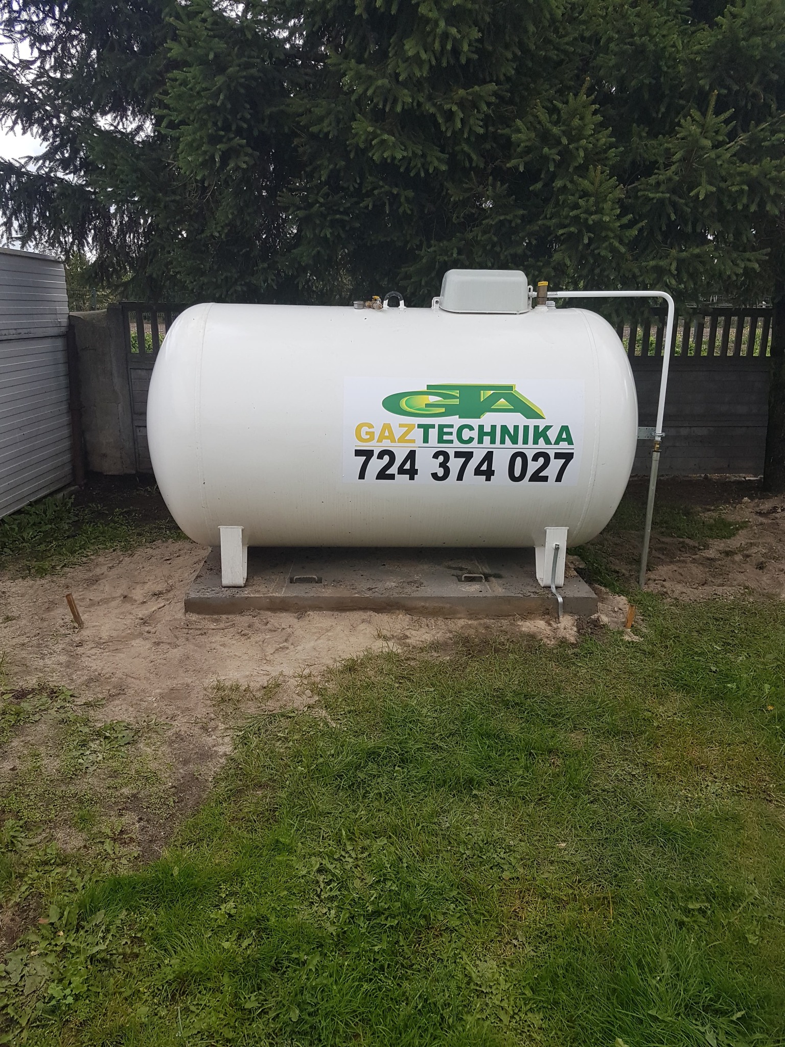 Biały zbiornik na gaz LPG z logo firmy Gaztechnika, umieszczony na betonowej podstawie w ogrodzie, widoczny przewód doprowadzający gaz.