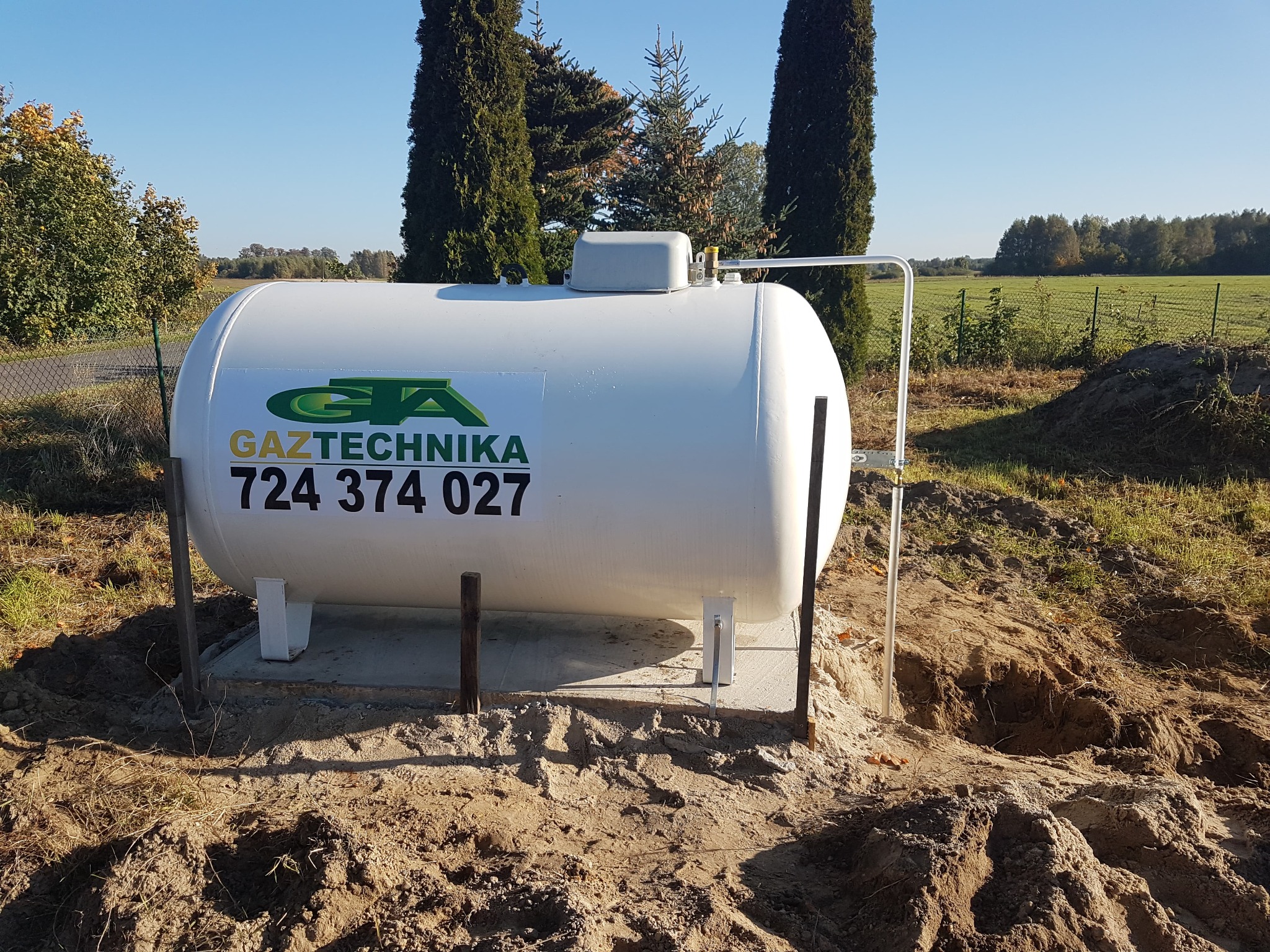 Biały, naziemny zbiornik gazu LPG z logo firmy Gaztechnika, osadzony na betonowej podstawie, otoczony wykopem w ziemi, na tle zieleni i błękitnego nieba.