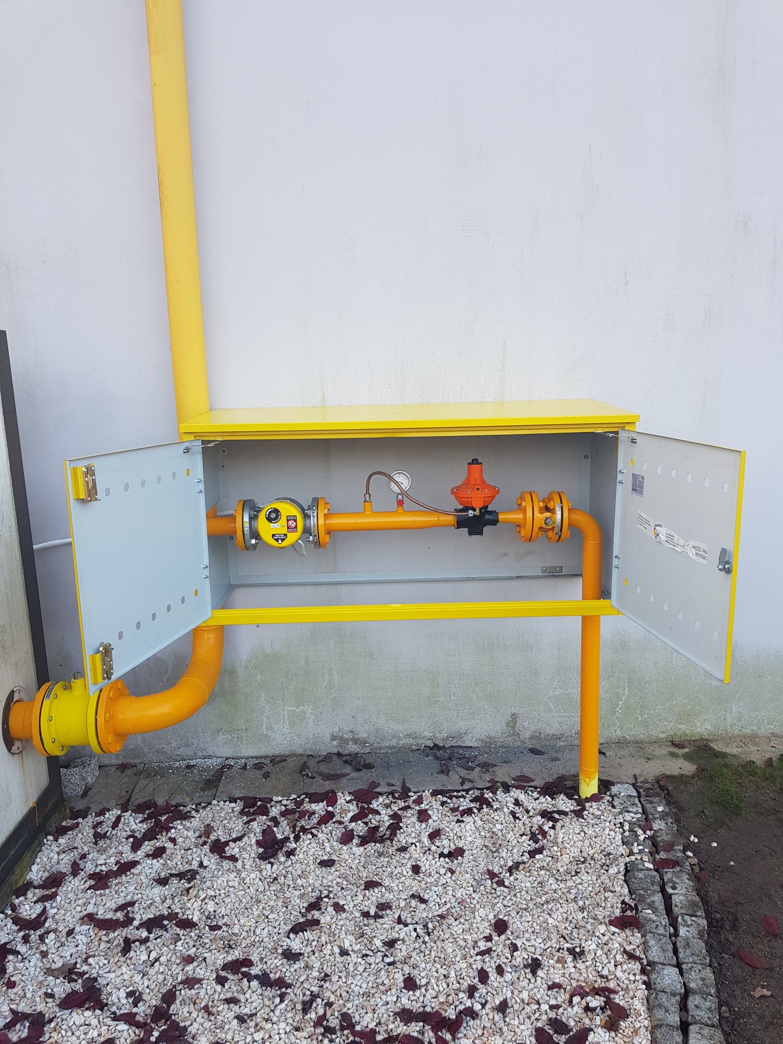 Żółta instalacja gazowa w skrzynce z otwartymi drzwiczkami, widoczny reduktor, licznik i rury na tle białej ściany i kamiennego podłoża.