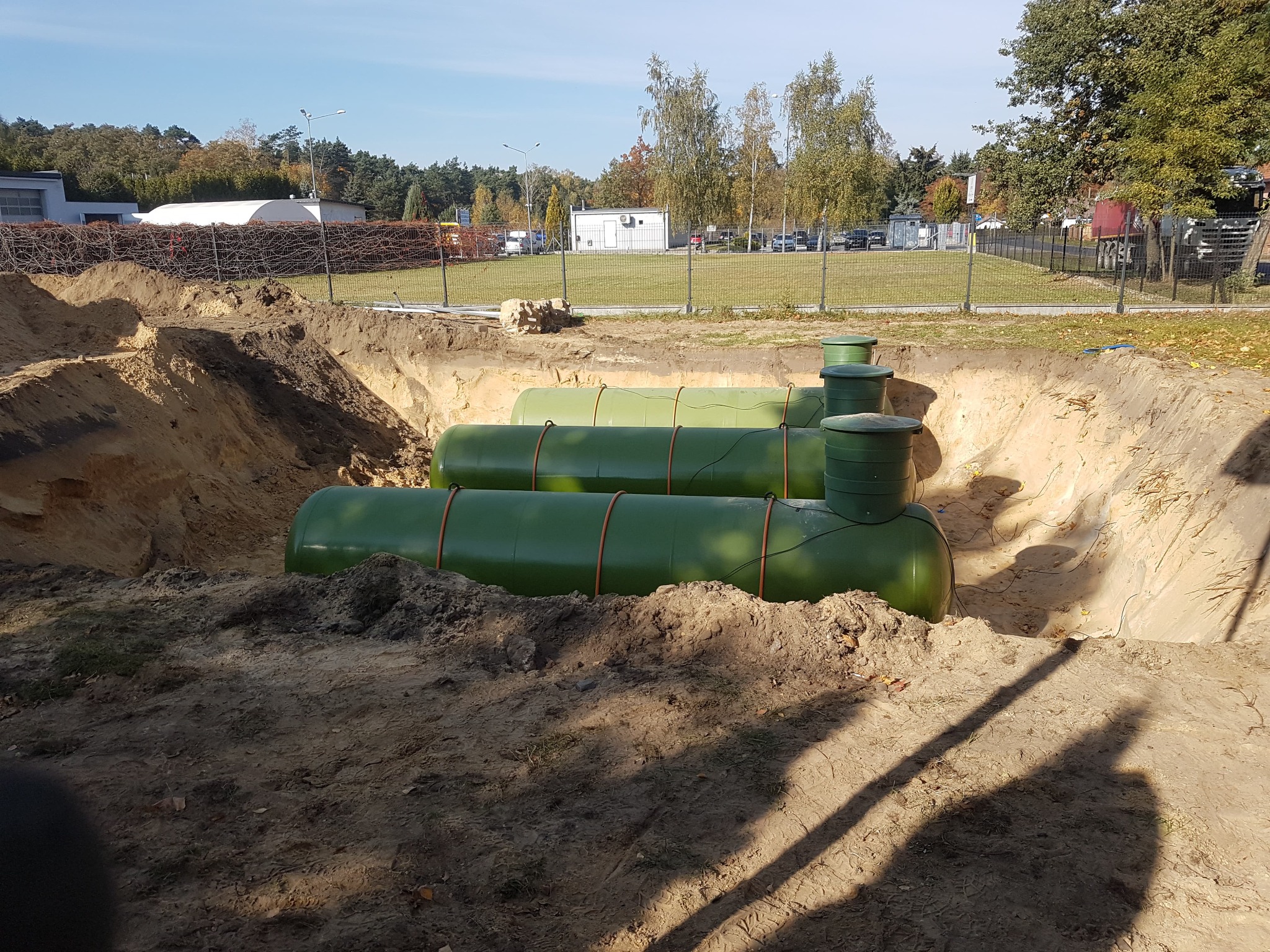 Trzy poziome, zielone zbiorniki w wykopie ziemnym, prawdopodobnie na gaz, z widocznymi włazami, na tle budynku i drzew