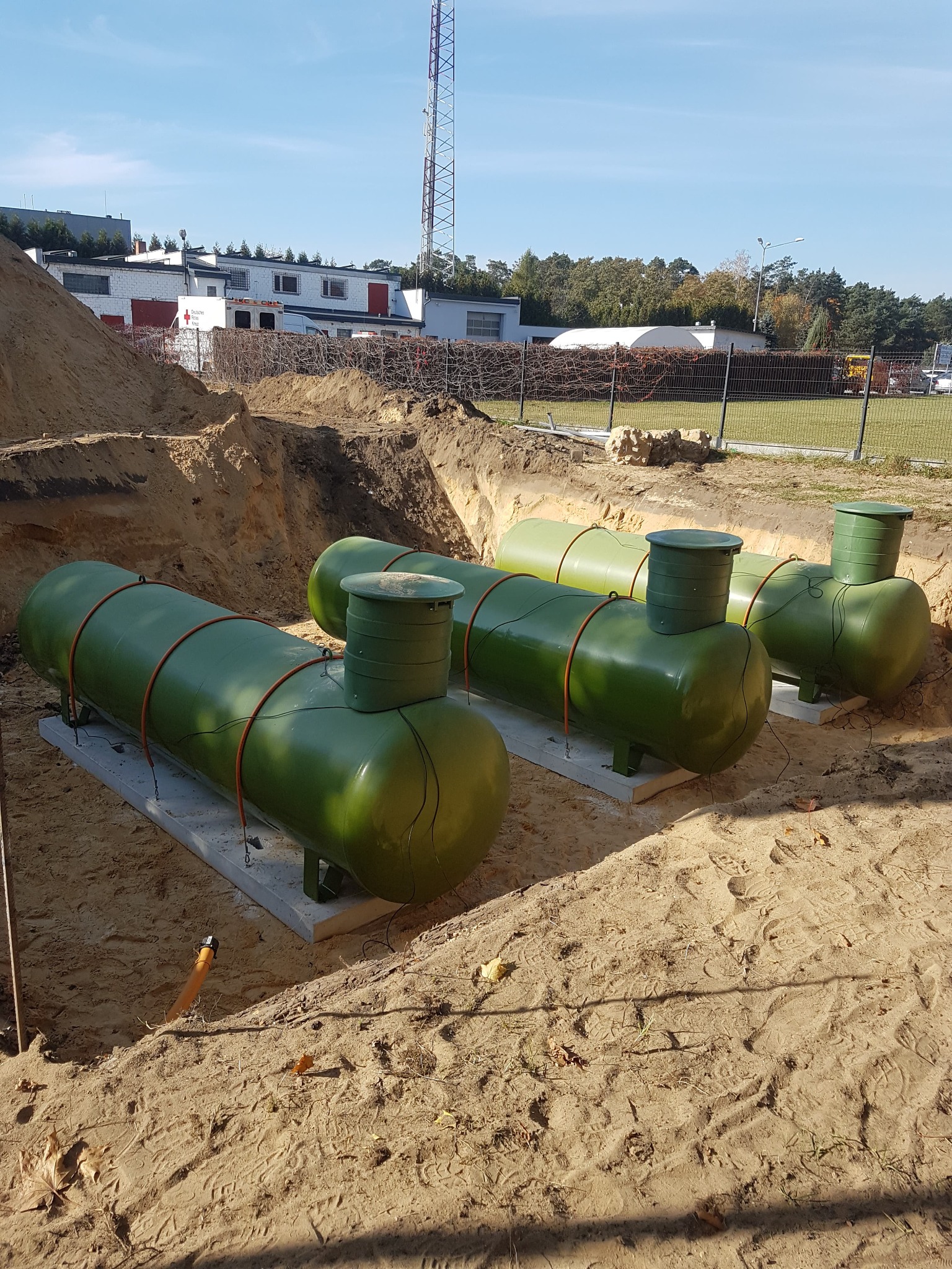 Zielone zbiorniki na gaz płynny, częściowo zagłębione w ziemi, umieszczone na betonowych fundamentach, z widocznym wykopem i rurą doprowadzającą, na tle budynku i wieży telekomunikacyjnej.