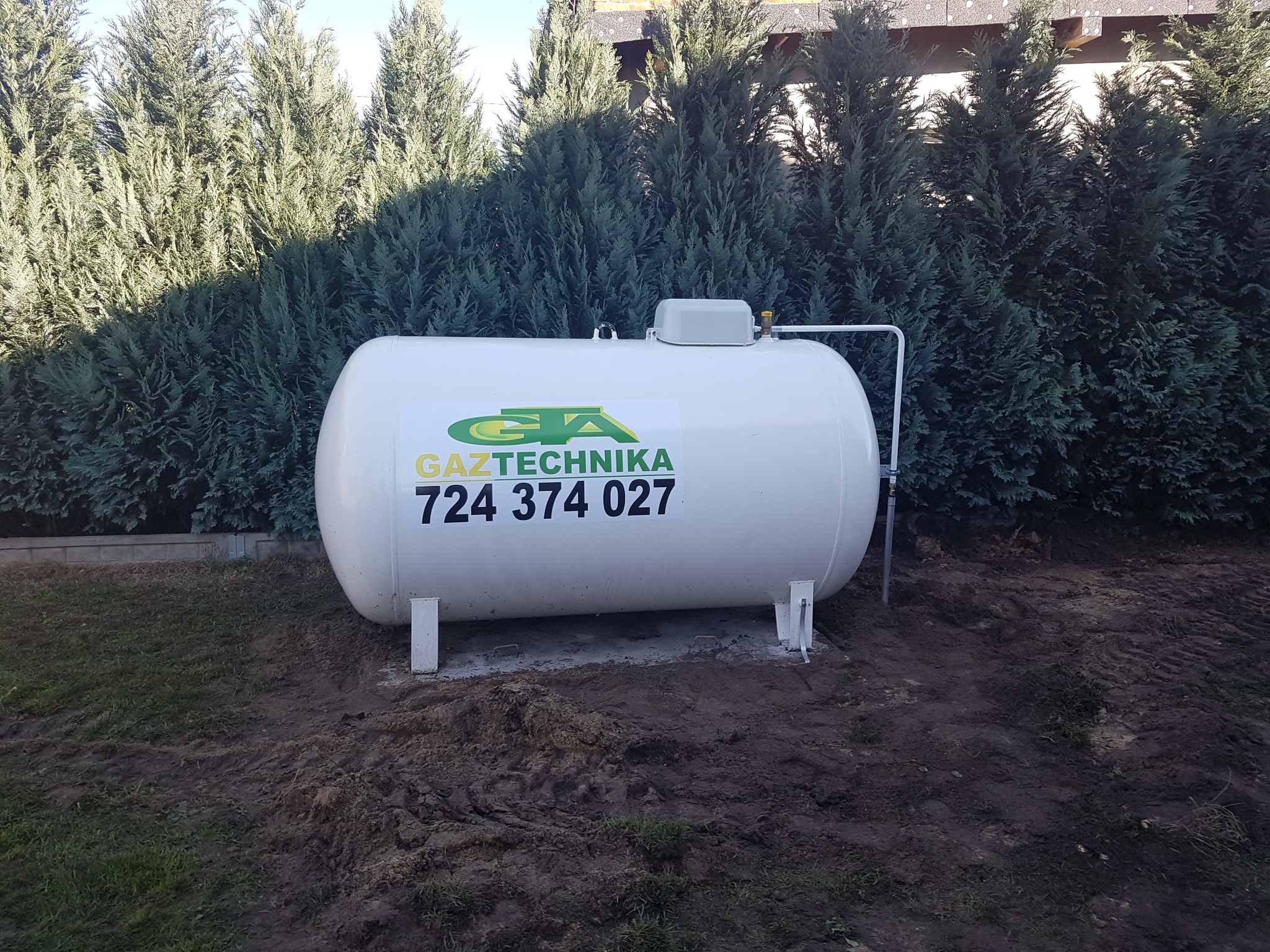 Biały zbiornik na gaz propan z naklejką firmy Gaztechnika, umieszczony na gruncie przed zielonymi tujami.