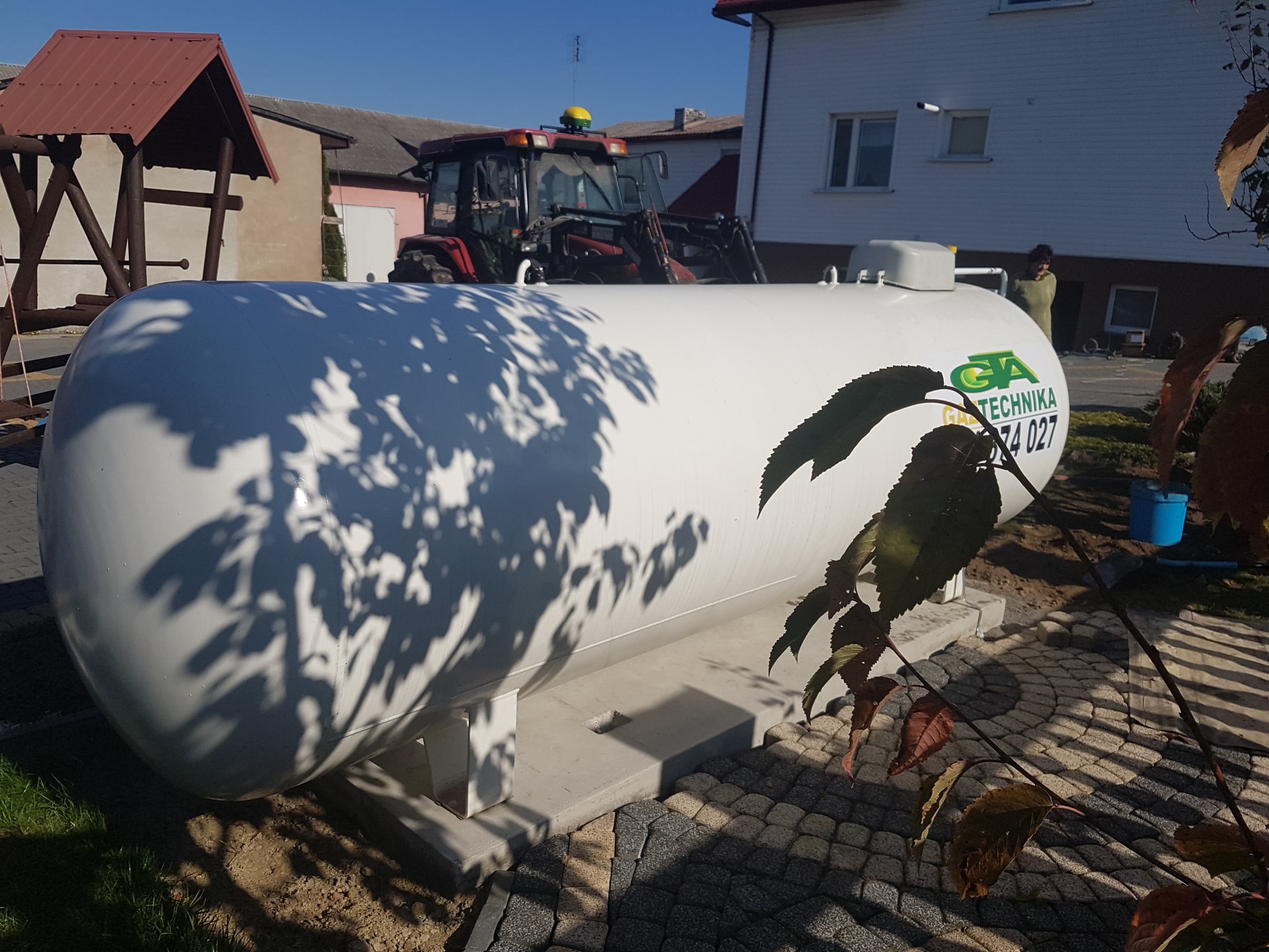 Biały zbiornik na gaz LPG z logo firmy, zamontowany na betonowej podstawie na posesji, w tle traktor i budynek mieszkalny.