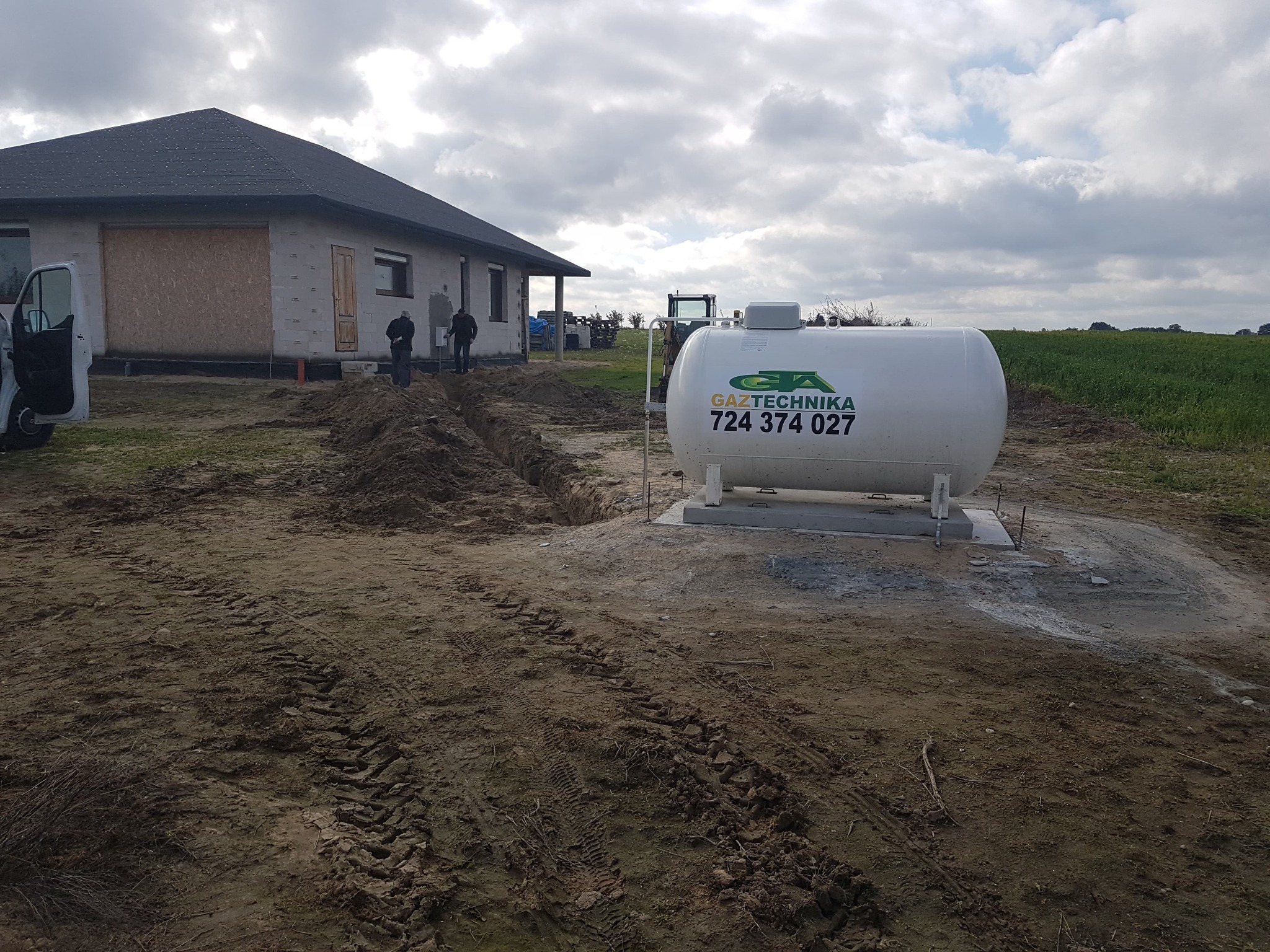Montaż naziemnego zbiornika na gaz płynny przy nowo budowanym domu jednorodzinnym, widoczny wykop pod instalację, logo firmy Gaztechnika i numer telefonu na zbiorniku.