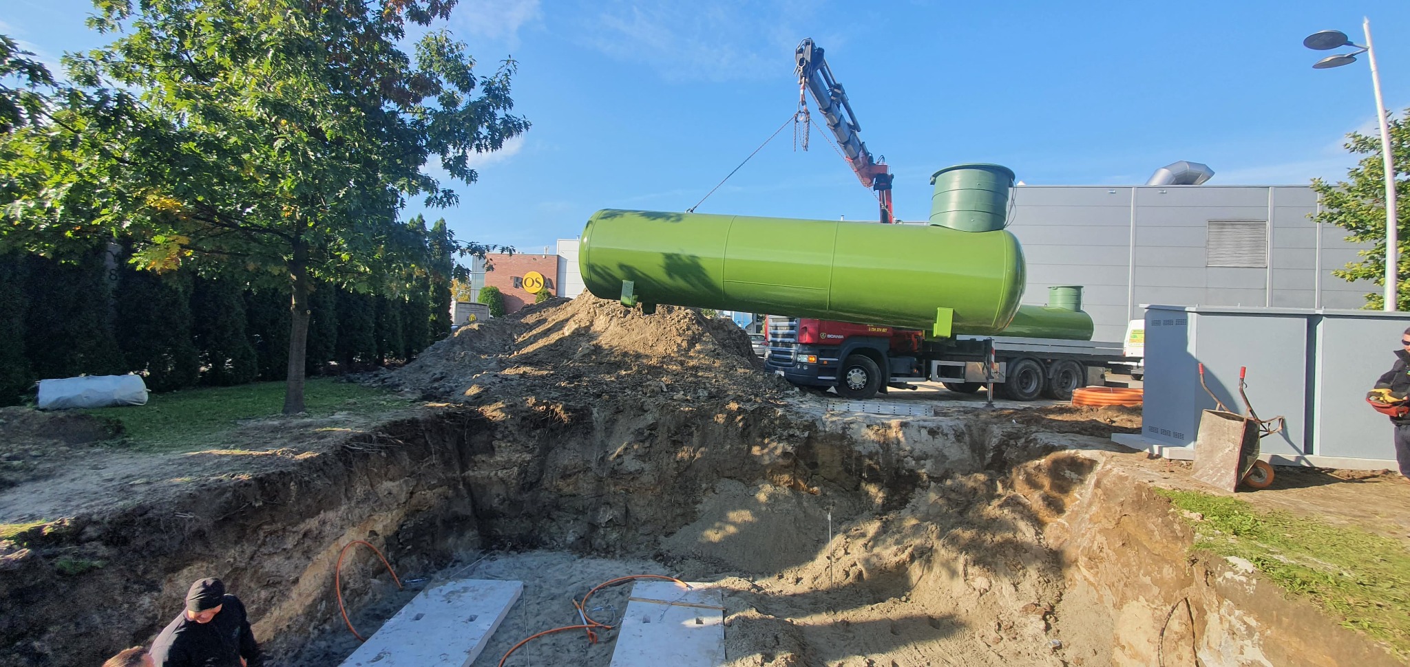 Montaż podziemnego zbiornika na gaz płynny za pomocą dźwigu, widoczny wykop z betonowymi płytami i pomarańczowymi rurami, ciężarówka z drugim zbiornikiem w tle.