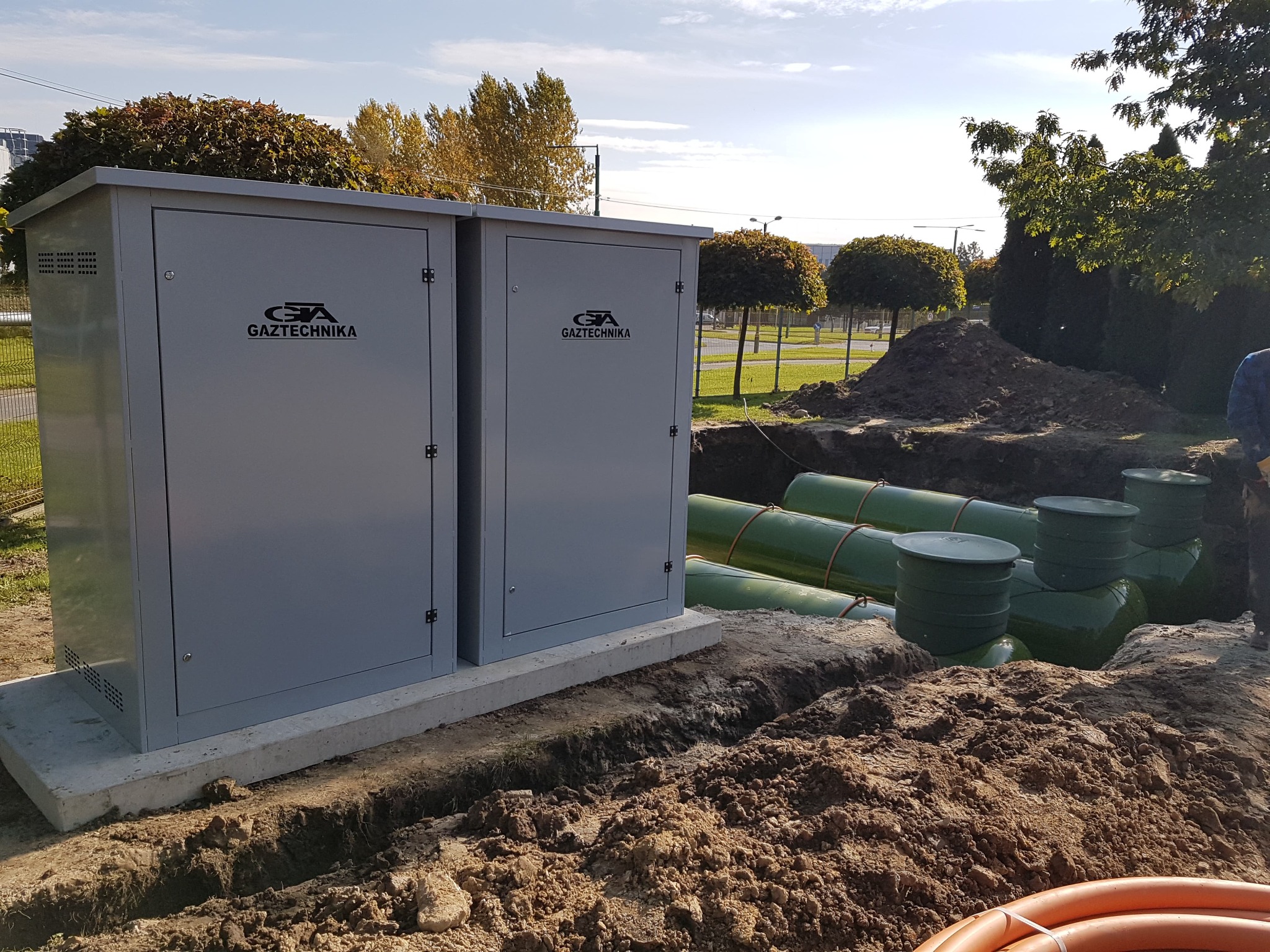Dwa szare, metalowe moduły z logo na betonowej podstawie, widoczne rury w wykopie, teren po robotach ziemnych, w tle drzewa i budynki.